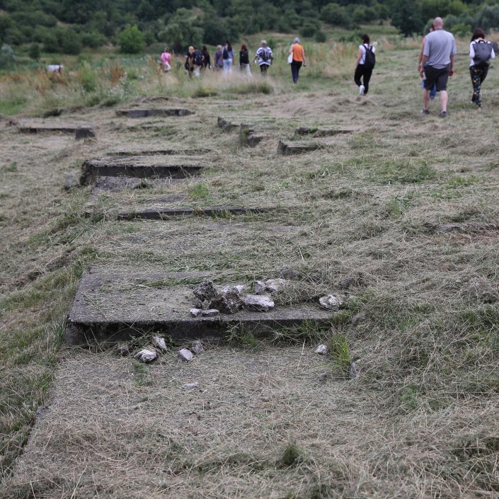 Na pierwszym planie widoczne pozostałości nagrobków otoczone bujną trawą. W dali grupa spacerujących ludzi.
