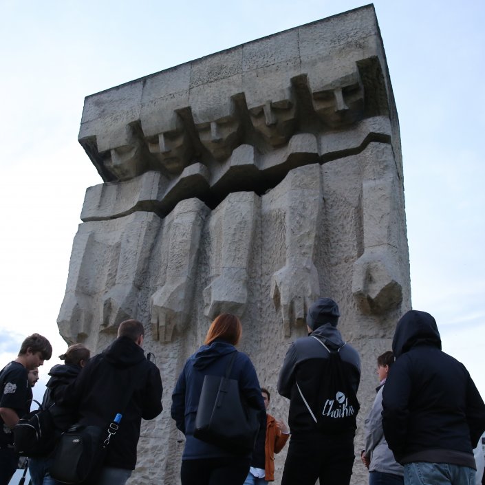 Duży wapienny pomnik przedstawiający pięć schematycznych postaci z wyrwą na wysokości piersi. Przed pomnikiem grupa ludzi.