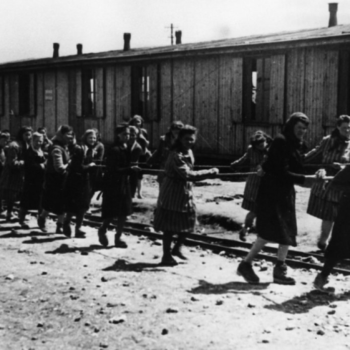 Fotografia archiwalna. Grupa więźniarek ciągnie metalowy wagonik wypełniony kamieniem. W tle drewniany barak.