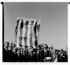 Zdjęcie archiwalne czarno-białe. Wysoki pomnik zasłonięty płachtą w ciemne i jasne pionowe pasy, przed nim widoczny tłum ludzi i starszy przemawiający człowiek.