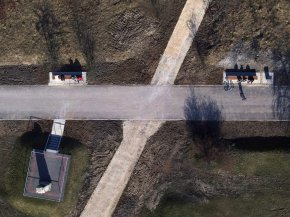 Zdjęcie z drona na dwie krzyżujące się drogi - asfaltową i kamienistą. Przy drugiej z nich znajudją się ławki, na który siedzą ludzie. U dołu widoczny obelisk