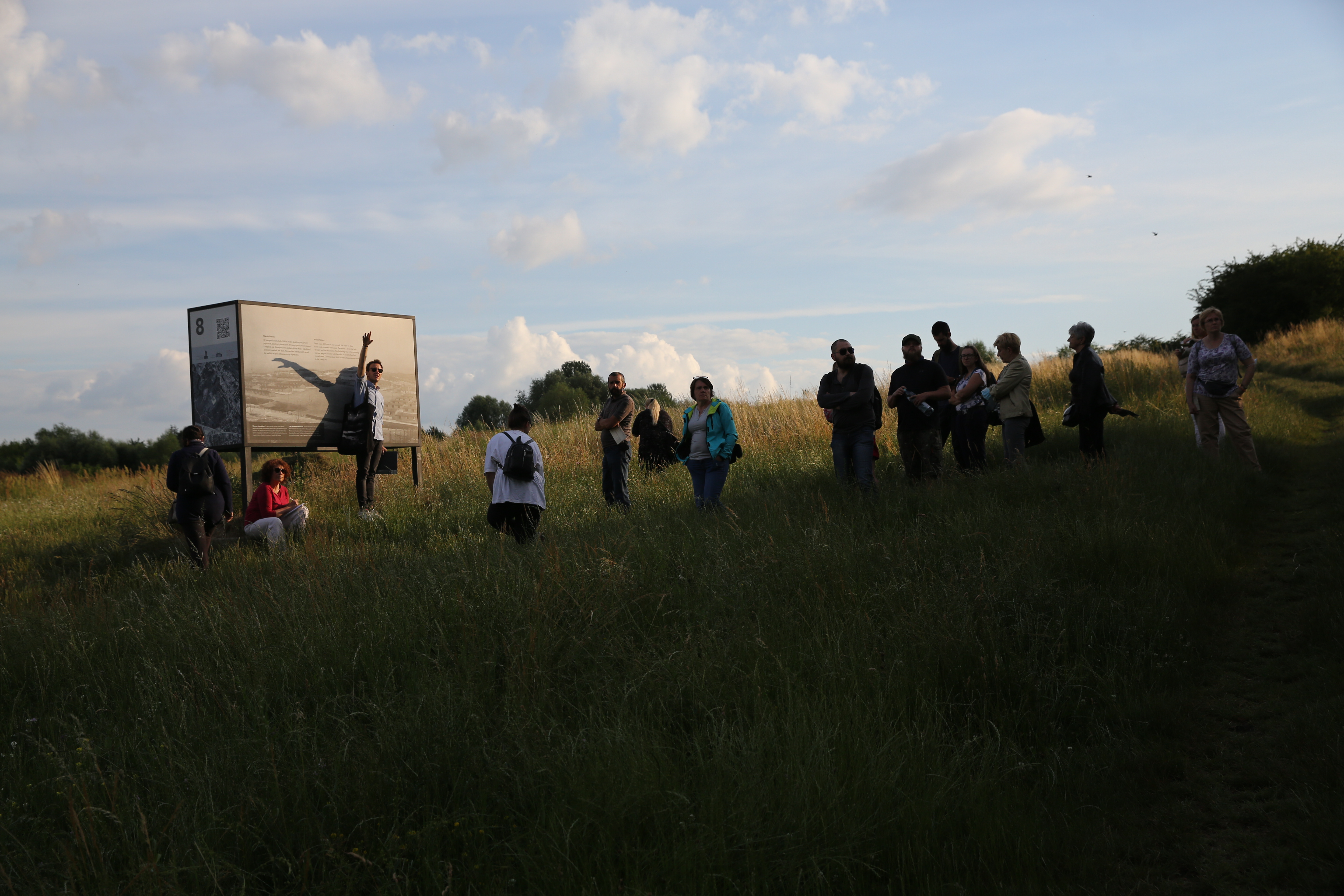 Grupa ludzi zwróconych w kierunku dużej tablicy z czarno-białym zdjęciem. Dookoła zielony krajobraz.