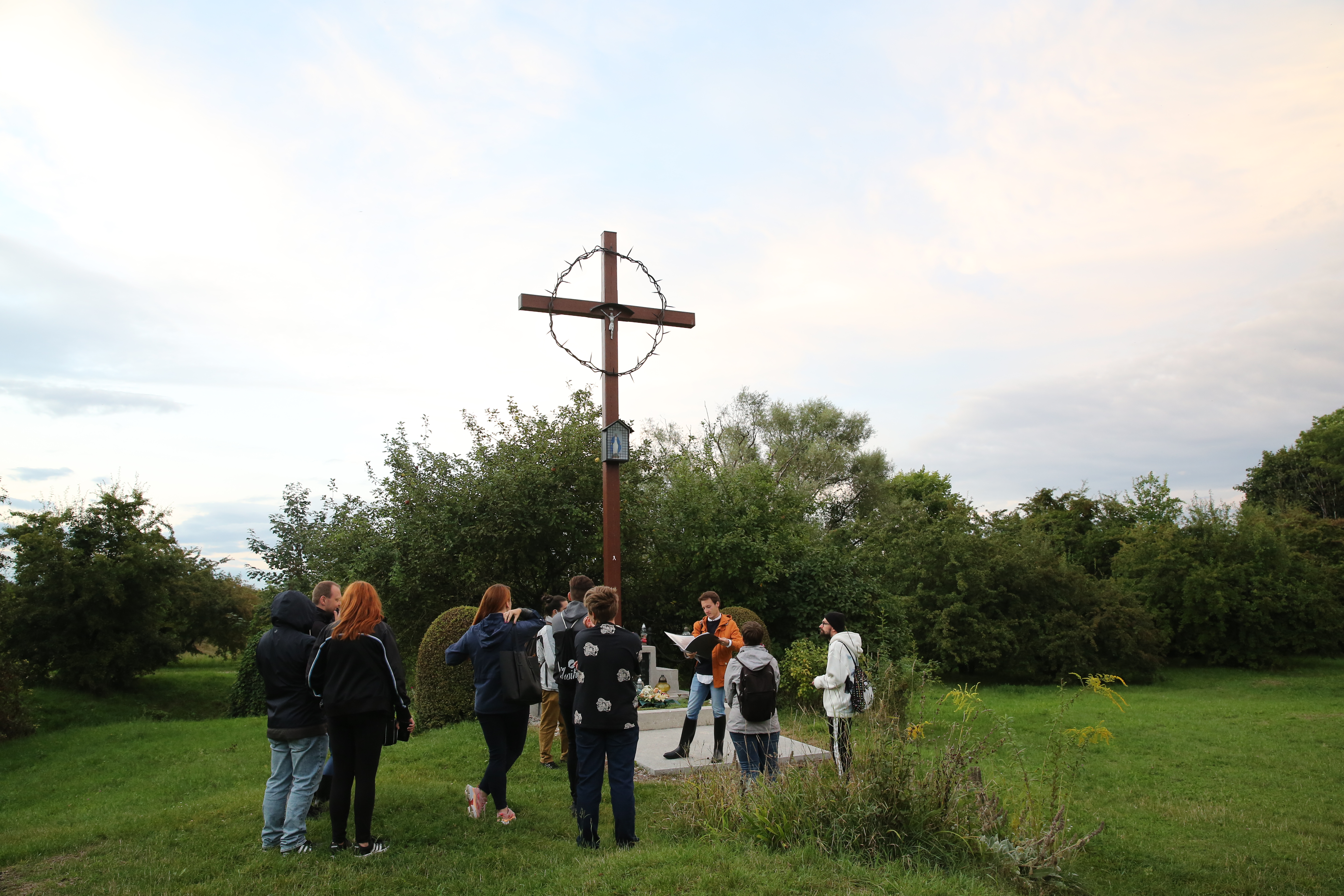Grupa ludzi zwróconych w kierunku krzyża na terenie KL Plaszow. Wokół zielona przestrzeń.