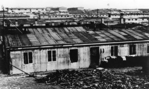 Zdjęcie archiwalne czarno-białe. Widok na długi parterowy barak z drewna. W tle liczne budynki.