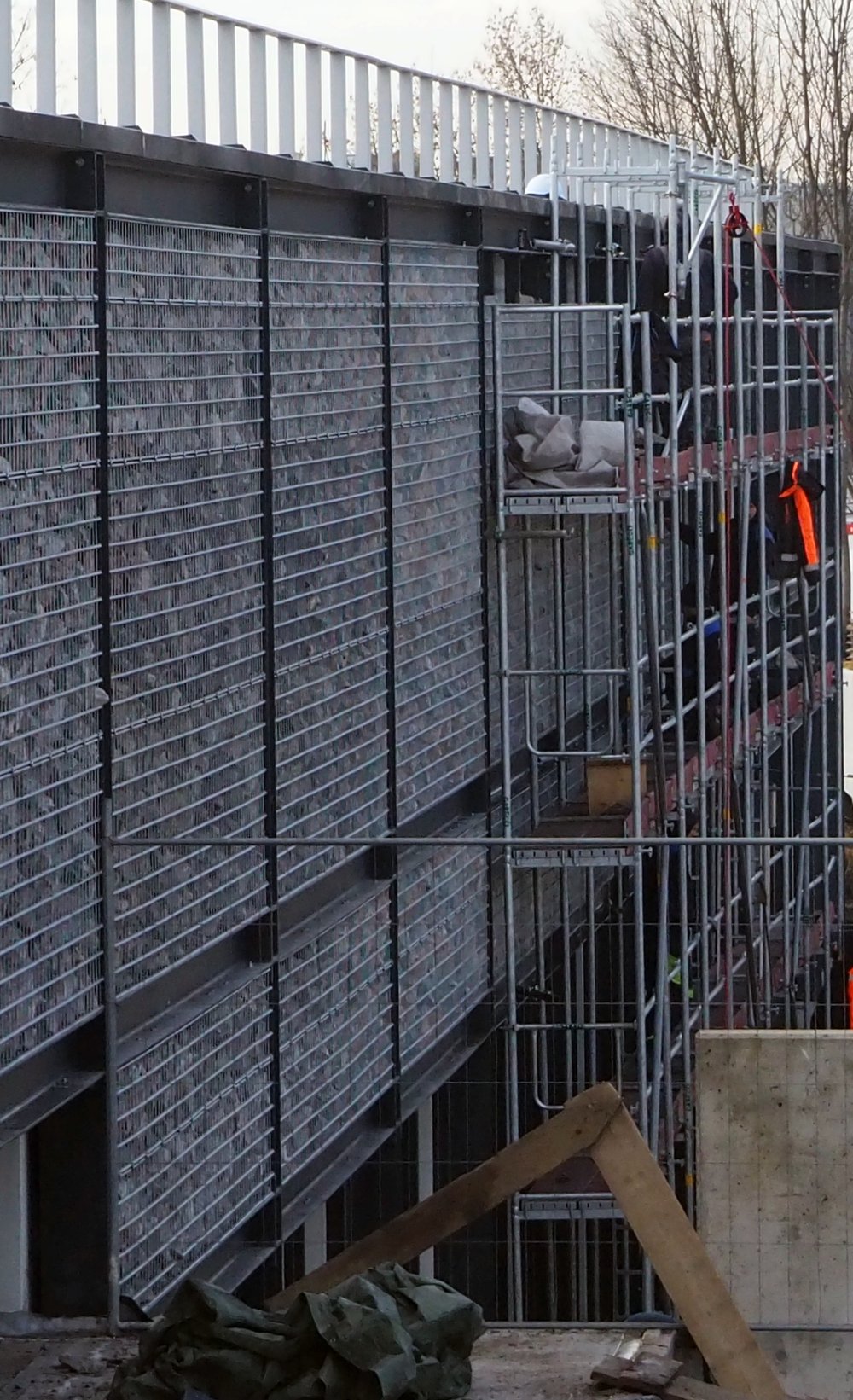 Widok na elewację budynku wykonanej w wapienny skał ułożonych w drucianych koszach. Przy budynku znajduje się rusztowanie, na którym pracują ludzie.